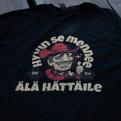 Musta Hyvin se mennee -t-paita, jossa cowboy-kuvitus ja teksti Älä hättäile, aseteltuna tasaiselle alustalle.