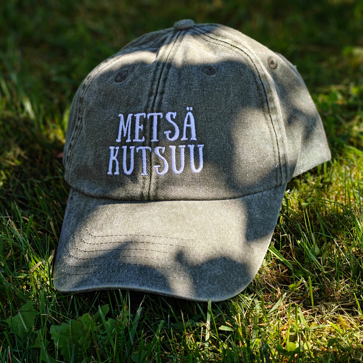 Vihreä Vintage Dad Hat -lippis Metsä kutsuu -kirjailulla, kuvattuna edestä sammalella.