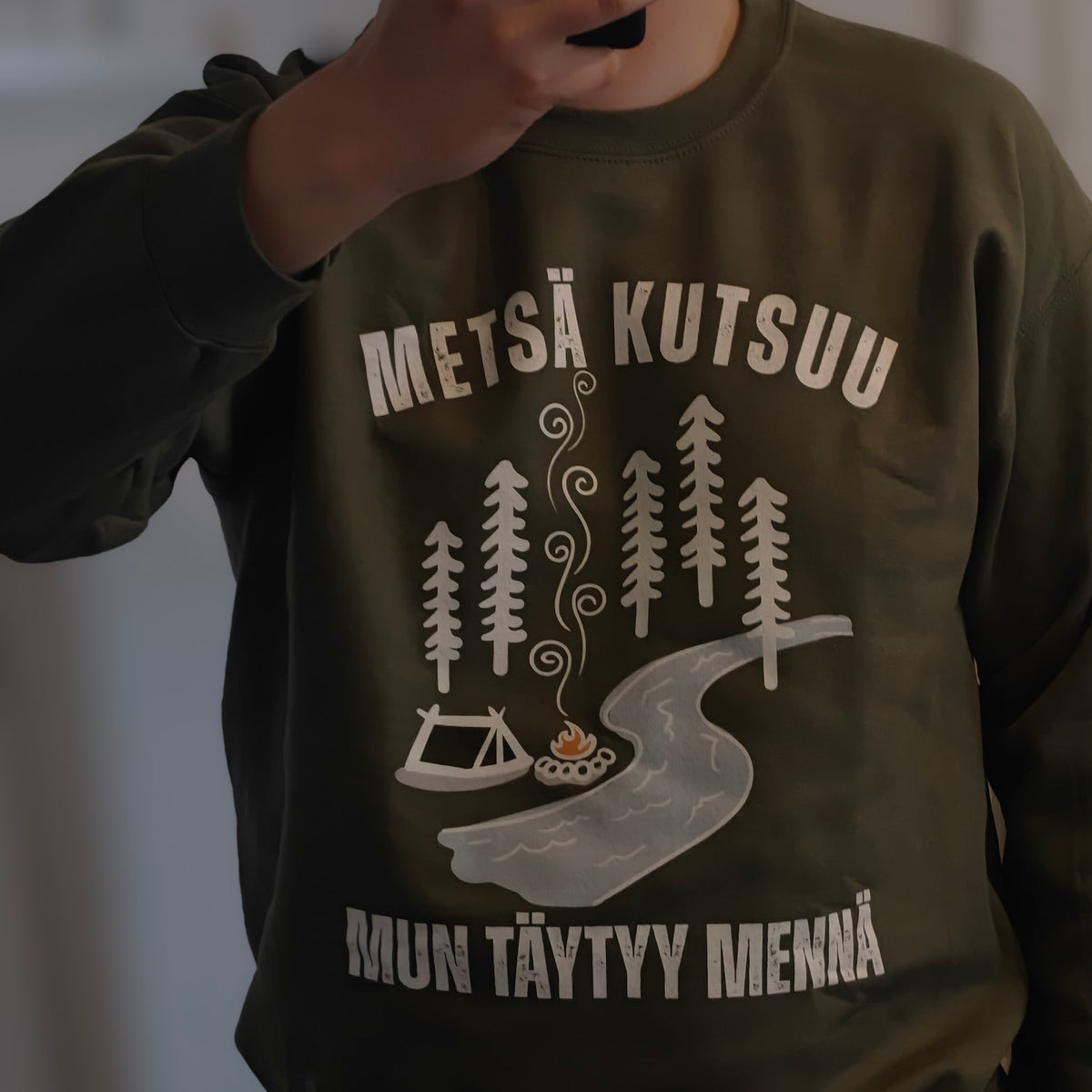 Vihreä Metsä kutsuu, mun täytyy mennä -collegepaita, jonka printissä on metsä, teltta, nuotio ja joki, henkilön päällä.